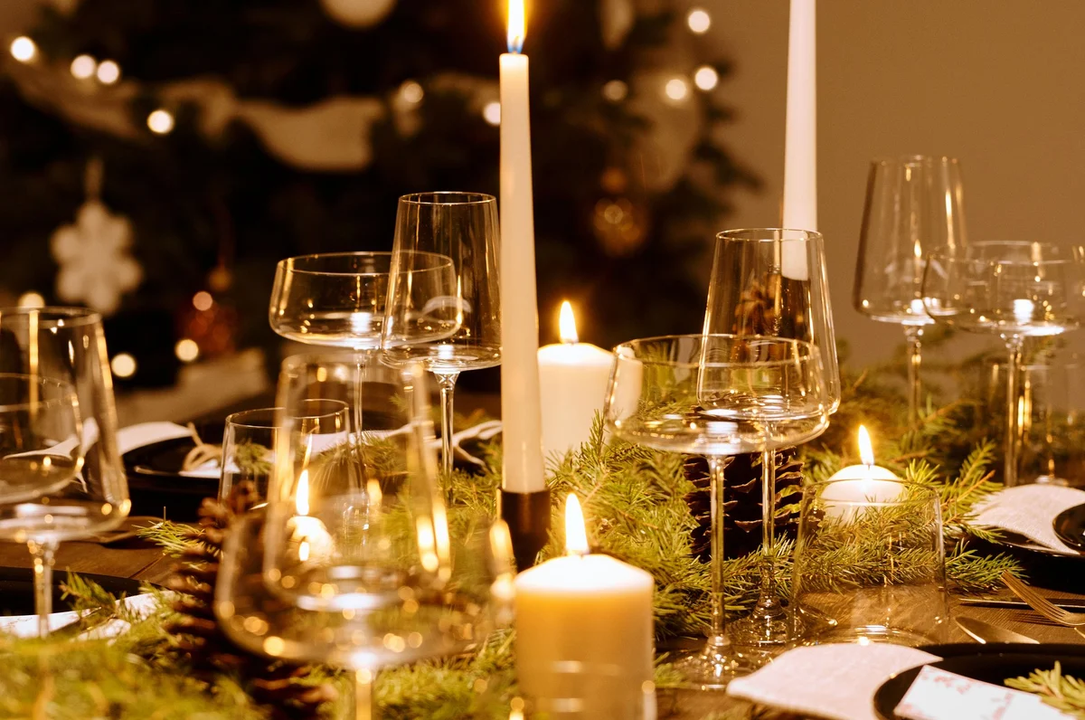Sfeerbeeld van een feestelijk gedekte kersttafel, met kaarslicht, dennengroen en glazen.